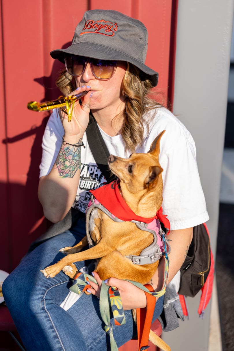 Woman in Bogey's hat and sunglasses blows a party horn, holding a small dog.