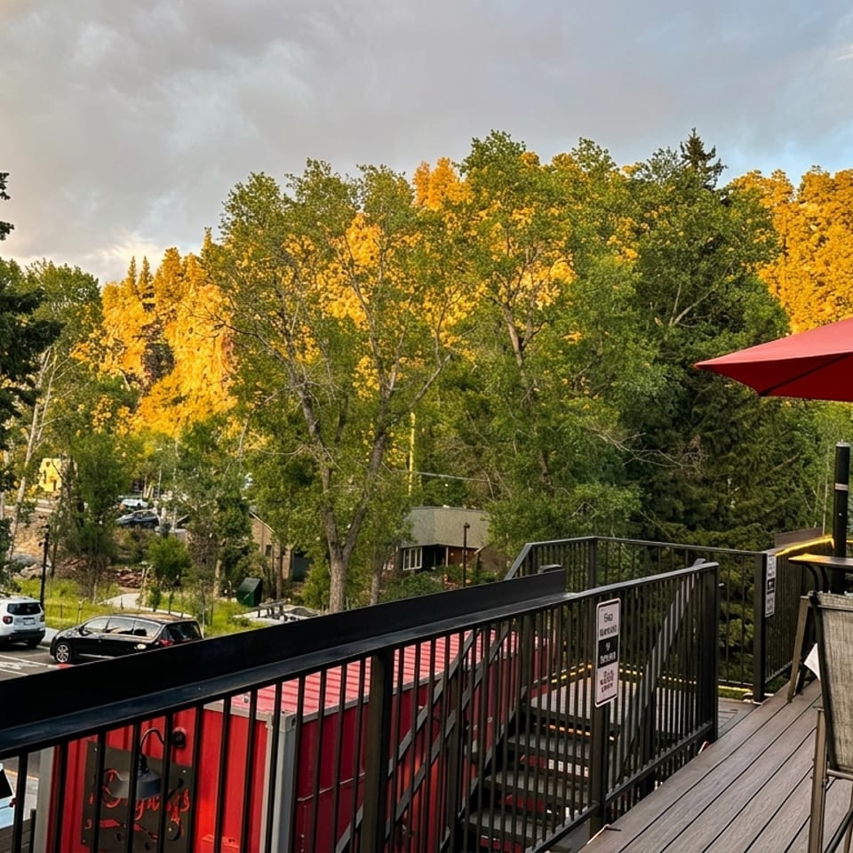 Outdoor restaurant patio with patrons under a red umbrella, overlooking a street and sunlit trees.