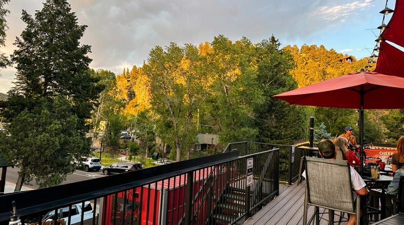 Outdoor restaurant patio with patrons under a red umbrella, overlooking a street and sunlit trees.