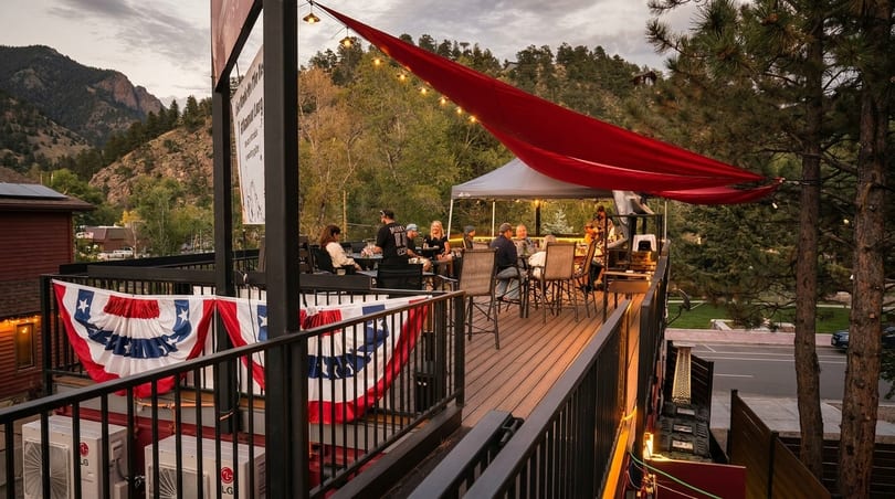 Patrons enjoying an outdoor restaurant patio with a red canopy, string lights, and mountain views.