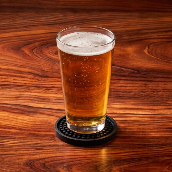 A tall glass of golden beer with a foamy head sits on a black coaster on a wooden table.