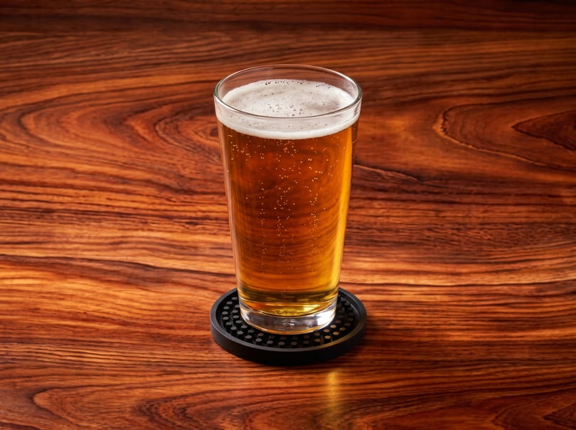 A tall glass of golden beer with a foamy head sits on a black coaster on a wooden table.
