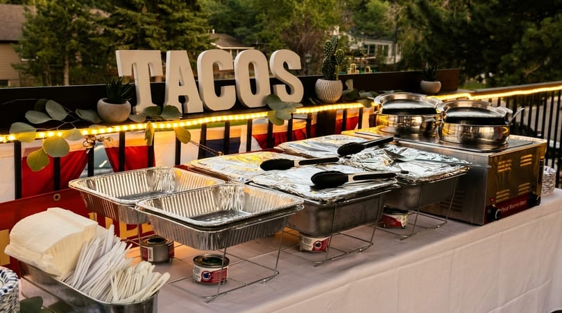 Outdoor taco bar with "TACOS" sign, chafing dishes, and serving utensils on a white tablecloth.