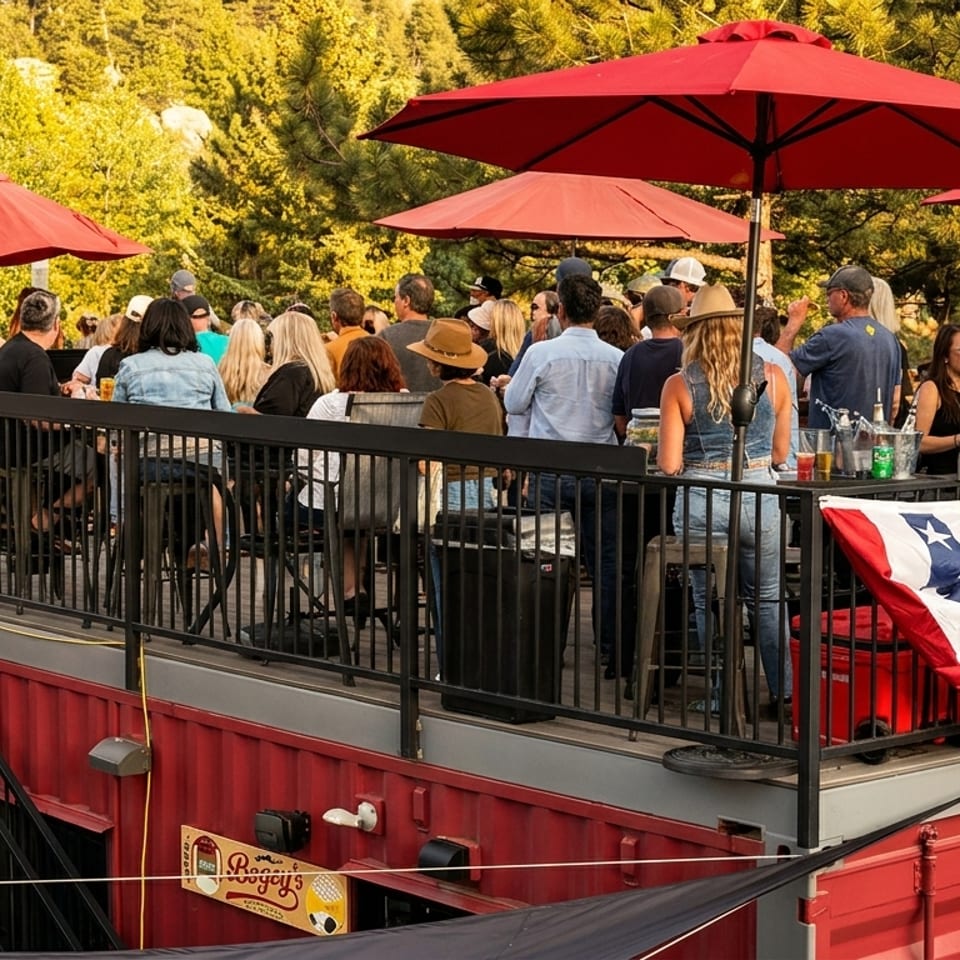 People socialize on a lively outdoor patio with red umbrellas and patriotic bunting, built on red shipping containers.
