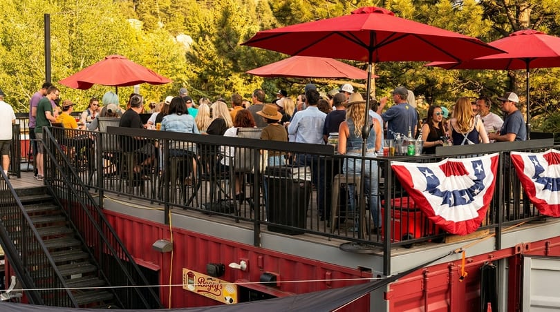 People socialize on a lively outdoor patio with red umbrellas and patriotic bunting, built on red shipping containers.