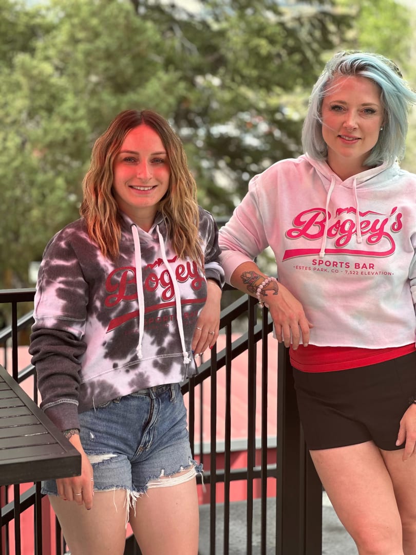 Two women wearing Bogey's Sports Bar hoodies pose outdoors on a patio with a railing.