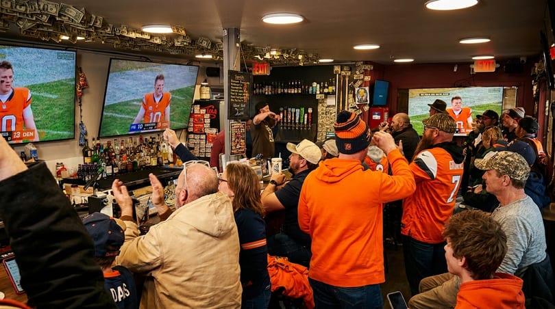 Patrons in orange and blue cheer while watching a football game on multiple TVs in a lively bar.
