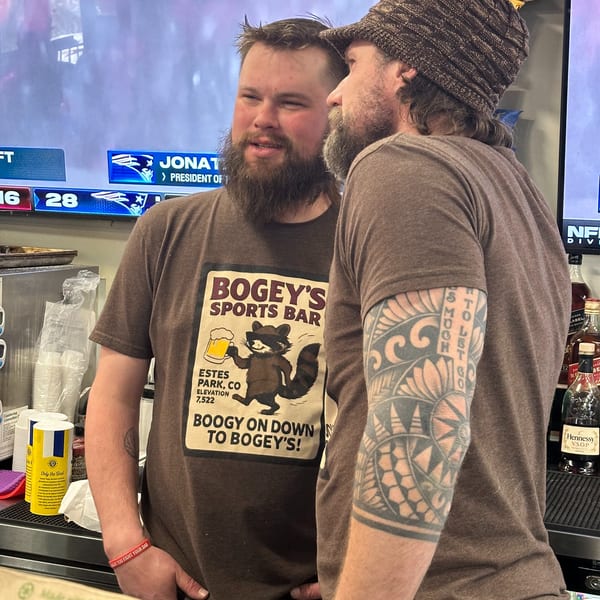 Two bearded men chat at a sports bar counter, one wearing a "Bogey's Sports Bar" t-shirt.