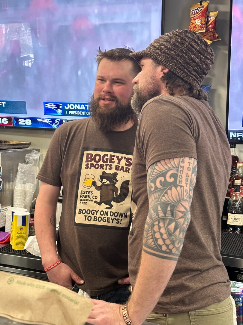 Two bearded men chat at a sports bar counter, one wearing a "Bogey's Sports Bar" t-shirt.