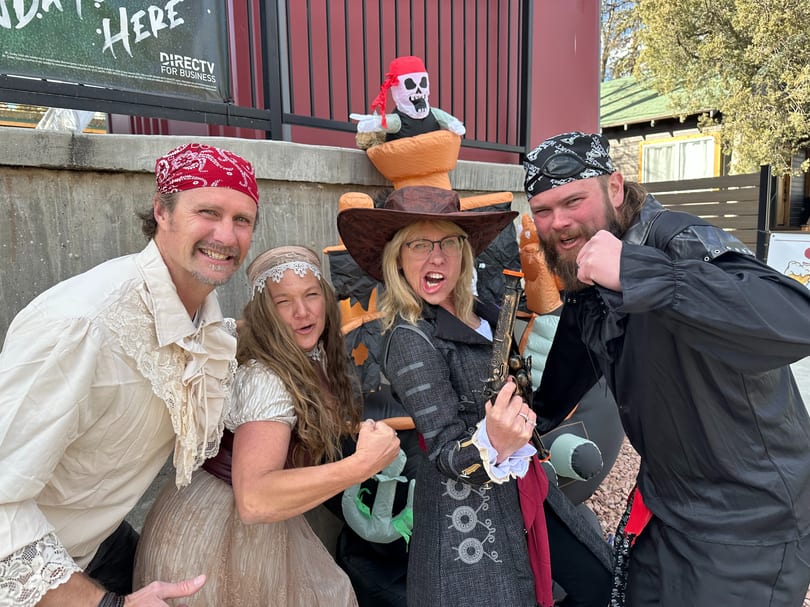 Four smiling adults in pirate costumes pose outdoors with a pirate skeleton inflatable.