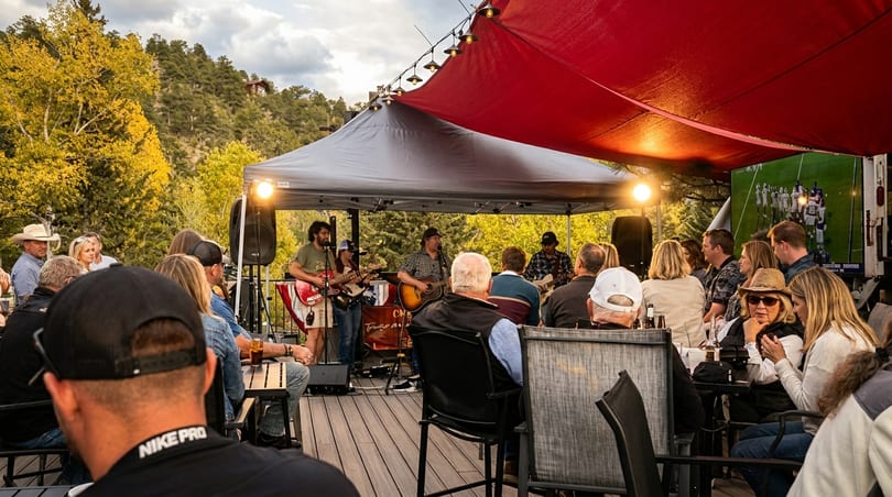 Patrons enjoy live music and a football game on an outdoor patio under red and gray awnings.