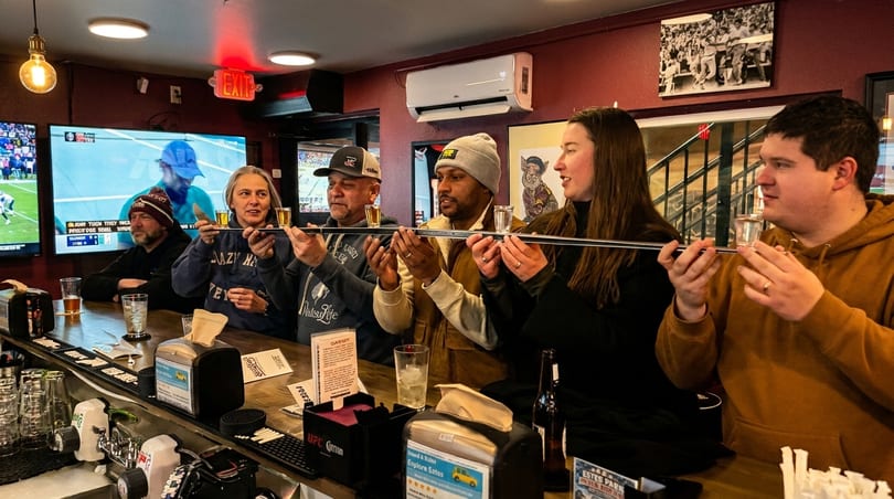 Six people at a bar counter participate in a group shot ski, with TVs showing sports in the background.