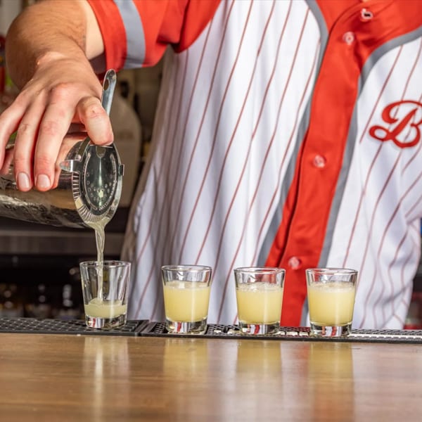 Bartender in a red and white striped shirt pouring yellow shots from a cocktail shaker.