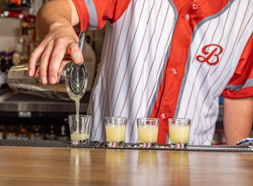 Bartender in a red and white striped shirt pouring yellow shots from a cocktail shaker.