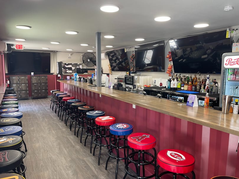 Interior view of a sports bar with a long counter, many team logo bar stools, and TVs.