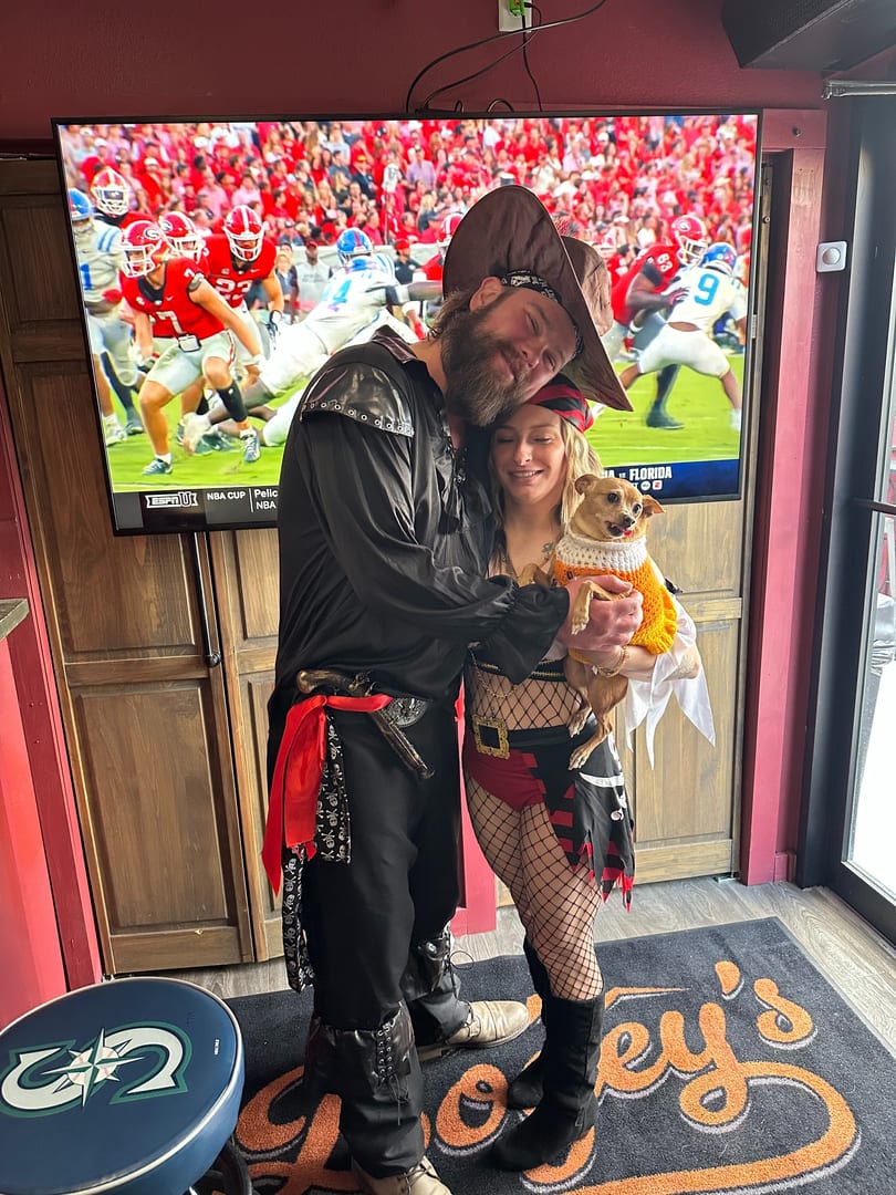 Pirate-costumed couple holds a dog in a sweater at Joey's, with a football game on a large TV.