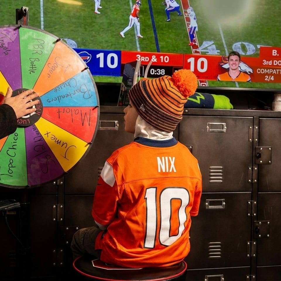A woman spins a prize wheel while a child in a Broncos jersey watches football on a TV.