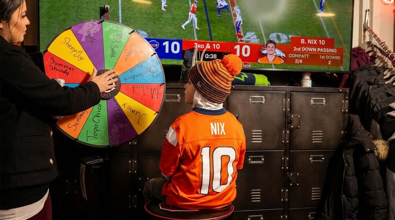 A woman spins a prize wheel while a child in a Broncos jersey watches football on a TV.