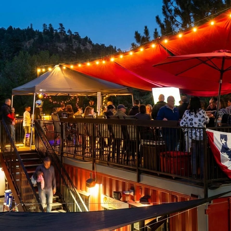 Outdoor restaurant patio at dusk with string lights, red awnings, and people socializing.