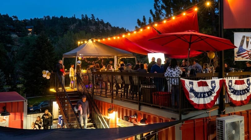 Outdoor restaurant patio at dusk with string lights, red awnings, and people socializing.