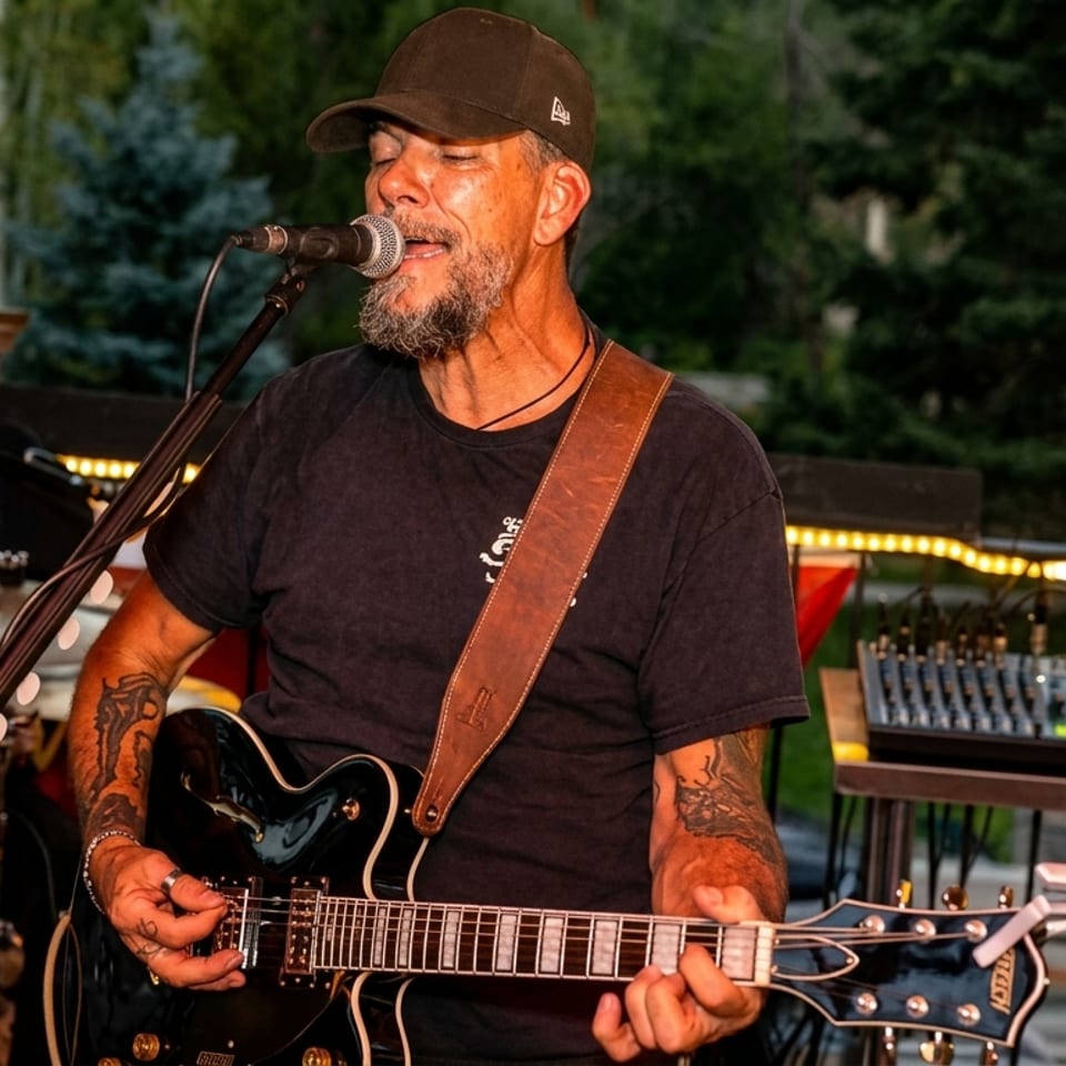 A man with a beard and cap sings into a microphone while playing an electric guitar outdoors.