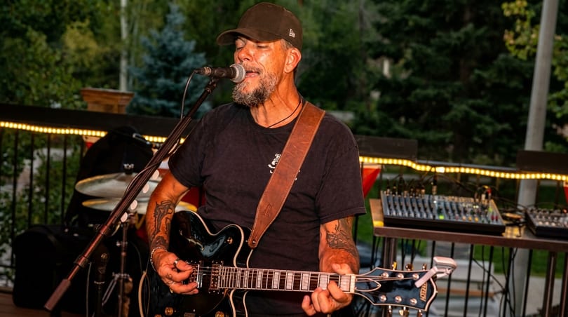 A man with a beard and cap sings into a microphone while playing an electric guitar outdoors.