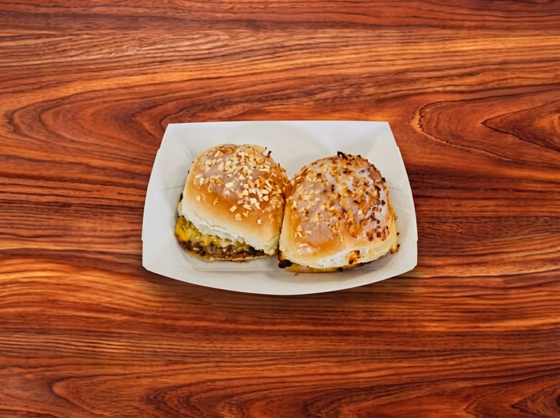 Two mini cheeseburgers with melted cheese and toasted buns in a white paper tray on a wooden table.