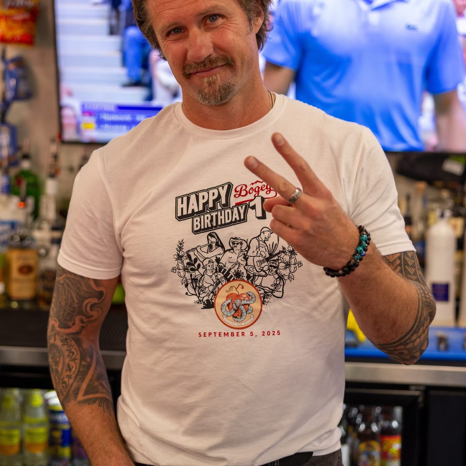 Man in a white 'Happy Birthday Bogey 1' t-shirt makes a peace sign, with a tennis match on a screen behind him.