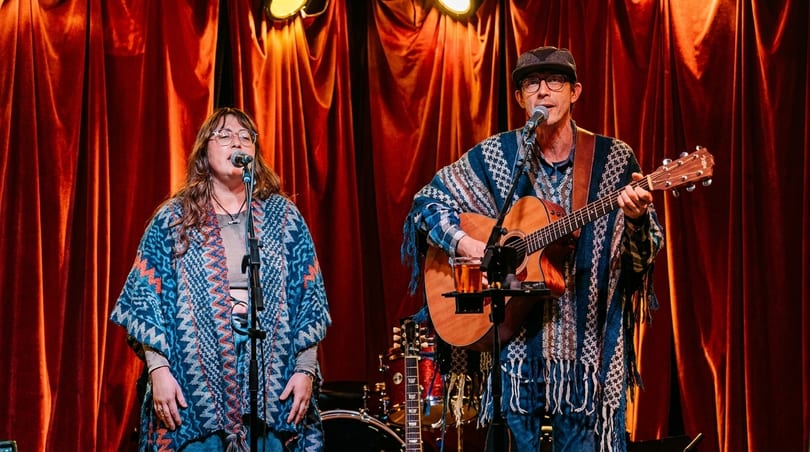 Two musicians, a woman and a man, perform on stage with microphones and a guitar.