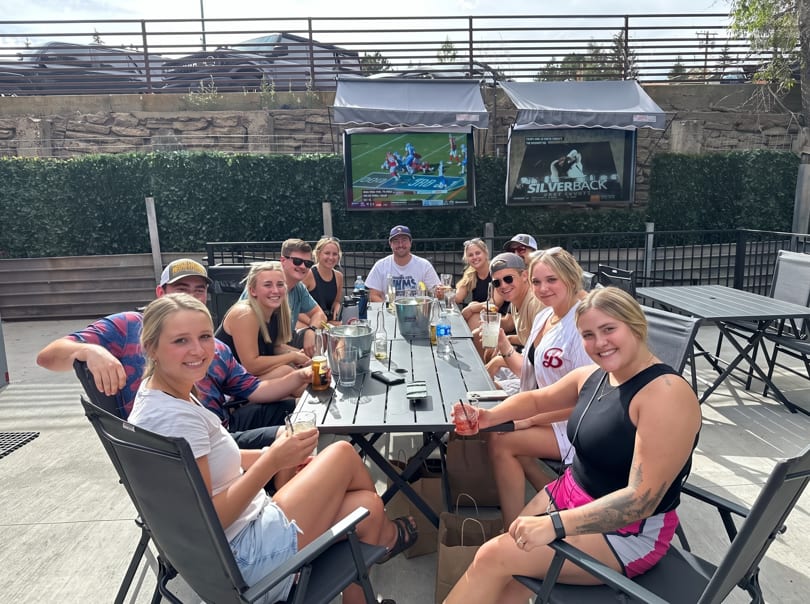 A group of friends enjoying drinks at an outdoor patio table with TVs showing sports.